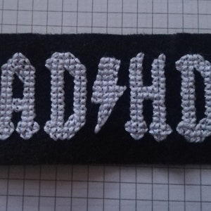 May include: Black rectangular patch with the word "ADHD" embroidered in white, gothic-style lettering. A white lightning bolt replaces the letter "I". The patch is on a gridded surface.