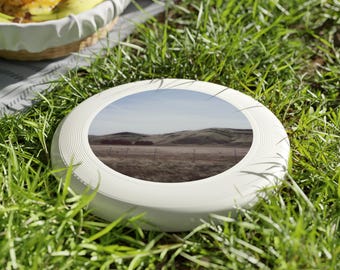 Può includere: Frisbee di plastica bianca con un'immagine stampata di un campo erboso e colline in lontananza.