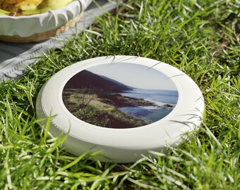 Può includere: Frisbee di plastica bianca con un'immagine stampata di un paesaggio costiero. L'immagine mostra una costa rocciosa con un oceano blu e una collina verde.