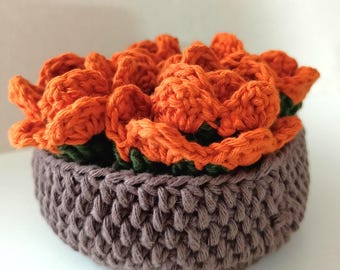May include: A handmade crochet succulent arrangement. The orange flowers sit in a brown crocheted basket. The flowers have green accents. The arrangement is a decorative item.