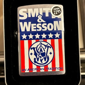 May include: A Smith & Wesson Zippo lighter in a black case. The lighter features a blue and white design with the words "SMITH & WESSON" and a series of white stars. The bottom half has red and white stripes with a circular logo and the text "AN AMERICAN TRADITION."