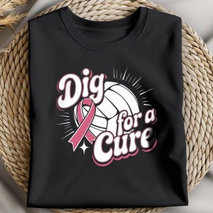May include: Black t-shirt featuring a volleyball design with a pink ribbon and the words "Dig for a Cure" in white and pink lettering. The shirt is folded and displayed on a woven surface.