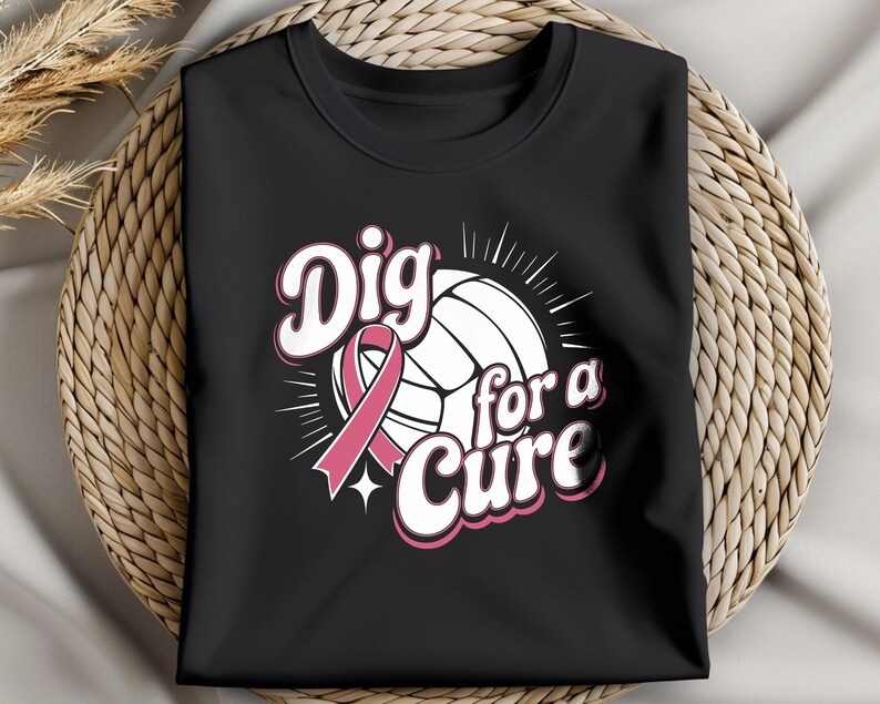 May include: Black t-shirt featuring a volleyball design with a pink ribbon and the words "Dig for a Cure" in white and pink lettering. The shirt is folded and displayed on a woven surface.