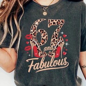 May include: A dark gray t-shirt with a leopard print design featuring the numbers "67" and the words "and fabulous". The numbers are decorated with red high heels.
