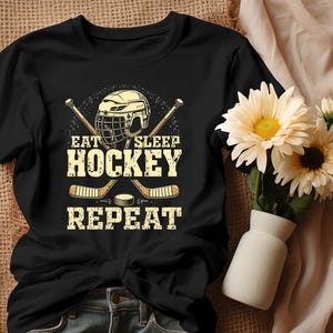 May include: Black t-shirt featuring a hockey helmet and sticks graphic. The text on the shirt reads "Eat Sleep Hockey Repeat."