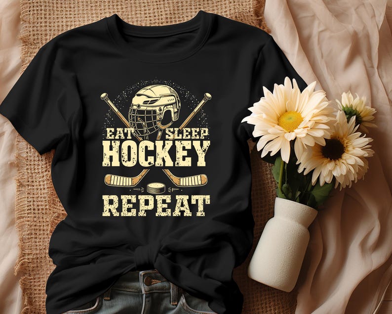 May include: Black t-shirt featuring a hockey helmet and sticks graphic. The text on the shirt reads "Eat Sleep Hockey Repeat."