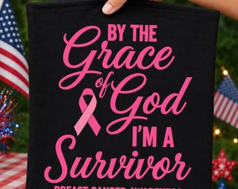 May include: A black fabric item featuring pink text that reads BY THE GRACE OF GOD I'M A SURVIVOR BREAST CANCER AWARENESS. The design includes a pink awareness ribbon graphic. The text is arranged in a mix of bold block and cursive fonts.