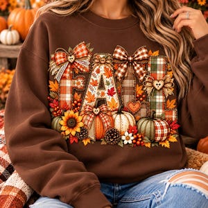 May include: A brown crewneck sweatshirt featuring a graphic print of the word FALL. The letters are decorated with plaid patterns, autumn leaves, pumpkins, sunflowers, pinecones, and decorative bows with small heart-shaped buttons.