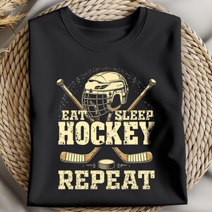 May include: Black t-shirt featuring a hockey helmet, sticks, and puck graphic. The text reads "Eat Sleep Hockey Repeat".