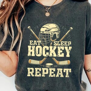May include: A dark grey t-shirt featuring a hockey helmet, sticks, and puck graphic. The text on the shirt reads "Eat Sleep Hockey Repeat".
