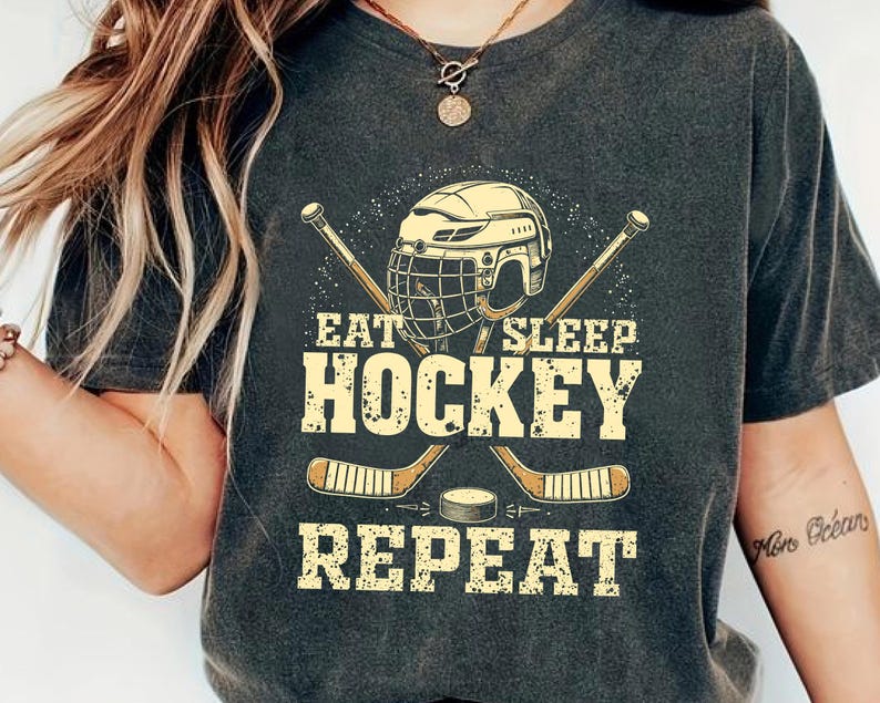 May include: A dark grey t-shirt featuring a hockey helmet, sticks, and puck graphic. The text on the shirt reads "Eat Sleep Hockey Repeat".