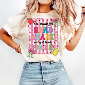 May include: Cream-colored t-shirt with the text "I'm ready for HEAD START but is it ready FOR ME?" in colorful letters. The design includes a red apple, pink bows, and a yellow notepad. The shirt is paired with blue jeans.