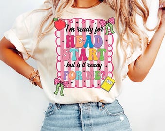 May include: Cream-colored t-shirt with the text "I'm ready for HEAD START but is it ready FOR ME?" in colorful letters. The design includes a red apple, pink bows, and a yellow notepad. The shirt is paired with blue jeans.