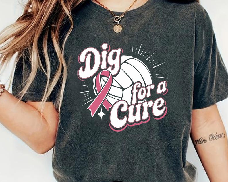 May include: Dark gray t-shirt with the words "Dig for a Cure" in white and pink lettering. A volleyball and pink ribbon are featured in the design. The shirt is a casual style.