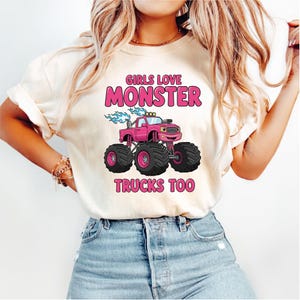 Può includere: T-shirt color crema con grafica di monster truck rosa e la scritta "GIRLS LOVE MONSTER TRUCKS TOO" in rosa. Il monster truck ha grandi pneumatici neri e fiamme blu.