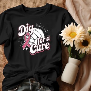 May include: Black t-shirt featuring a volleyball with a pink ribbon and the text "Dig for a Cure" in a pink and white design. The shirt is a crew neck style and is knotted at the waist. A vase of flowers is in the background.