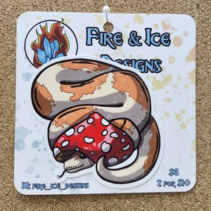 May include: A sticker featuring a cartoon snake coiled around a red mushroom with white spots. The sticker is on a white card with the text "Fire & Ice Designs" and a logo of fire and ice. The sticker is on a corkboard.