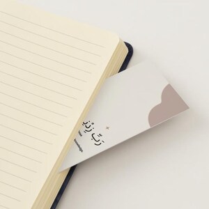 May include: A white notebook with lined pages and a blue spine. A bookmark with a pink cloud design and the text "تُرَتِّبُ" is sticking out of the notebook.