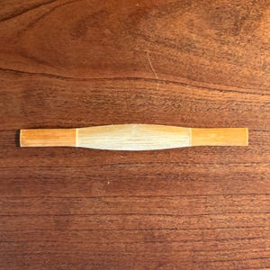 May include: A light brown, cylindrical bamboo object with a lighter, central section. The ends are a darker shade of brown. The object is lying on a wooden surface.