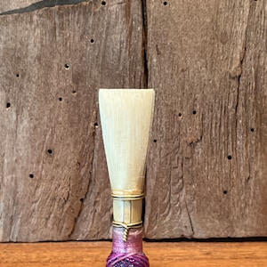 May include: A white wooden oboe reed with a purple and blue braided knot at the bottom.