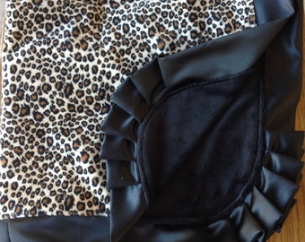 May include: A blanket featuring a leopard print pattern on one side and a solid black plush fabric on the other. The blanket is finished with a wide, pleated black satin ruffle border that frames the edge of the black fabric section.