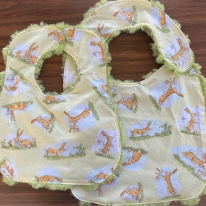 May include: Two yellow baby bibs with green trim and a brown rabbit pattern. The bibs are made of soft fabric and have a ruffled edge.