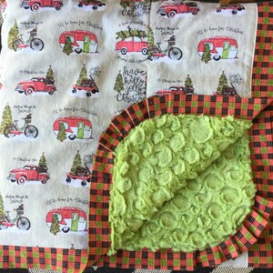 May include: A green and white Christmas-themed baby blanket with a red and black plaid border. The blanket features a variety of Christmas-themed illustrations, including a camper, a car, and a bicycle. The text "Have a holly jolly Christmas" is printed on the blanket.