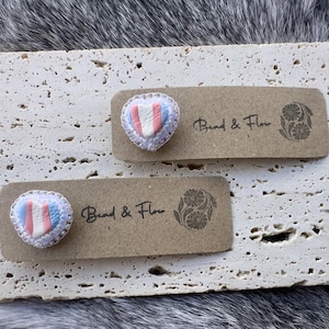 Peut inclure: Deux épingles en forme de cœur avec un motif rayé rose, blanc et bleu, encadrées de dentelle blanche. Elles sont fixées sur de petites étiquettes rectangulaires marron avec le texte "Bend & Flow" et un motif floral. Les épingles reposent sur une surface texturée blanc cassé.