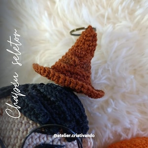 May include: A small, crocheted brown witch hat keychain with a bronze-colored ring. The hat is detailed with a pointed crown and a wide brim. The text "Chapéu Seletor" is written in white cursive on the left side of the image. The background is a fluffy white surface.