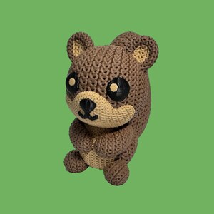 May include: A brown bear figurine with a knitted texture. The bear has black eyes, a tan muzzle, and tan inner ears. The bear is holding its paws together, and the background is green.