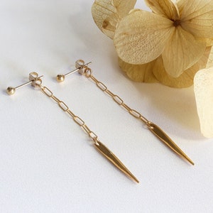May include: A pair of gold earrings with a delicate chain and a sharp, pointed pendant. The earrings are made of metal and have a simple, minimalist design.