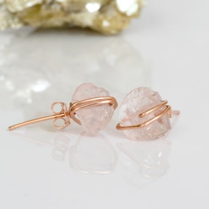 May include: A pair of rose quartz earrings with gold wire wrapping. The earrings are made with natural, raw rose quartz crystals. The rose quartz is a light pink color.
