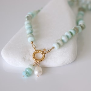 May include: A beaded necklace featuring light blue and gray stones, connected by gold-colored clasps. A gold ring clasp holds a light blue stone and a white pearl pendant. The necklace is displayed on a white surface.