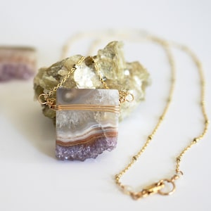 Puede incluir: Un collar de cadena dorada con un colgante de amatista y ágata envuelto en alambre. El colgante es un cristal natural en bruto con bandas moradas y blancas.