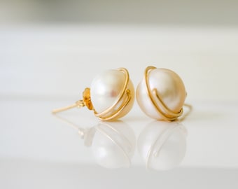 May include: A pair of stud earrings featuring round white pearls encased in thin gold wire frames. The earrings have gold posts and butterfly backs, with the pearls reflecting clearly on the smooth surface beneath them.