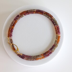May include: A multi-strand necklace with faceted beads in shades of orange, brown, and purple. The necklace is secured with a gold-coloured clasp. The beads are arranged in a circular pattern on a white surface.