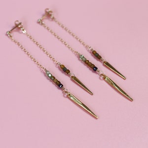 May include: A pair of gold earrings with a long chain and a gold spike at the end. The chain is adorned with small brown, green, and black beads.