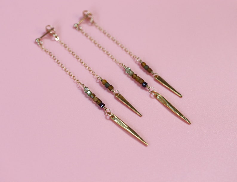 May include: A pair of gold earrings with a long chain and a gold spike at the end. The chain is adorned with small brown, green, and black beads.