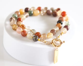 May include: A multi-colored beaded necklace with a gold clasp and a small rectangular charm. The beads are various colors, including red, orange, yellow, green, and brown. The necklace is displayed on a white surface.
