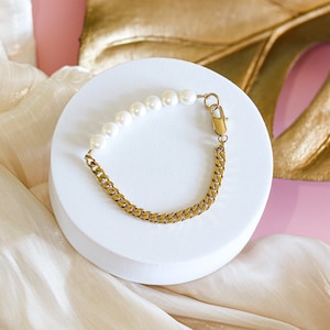 May include: A gold chain bracelet with a row of white pearls. The bracelet is on a white surface.