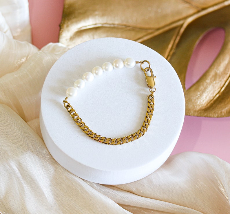 May include: A gold chain bracelet with a row of white pearls. The bracelet is on a white surface.