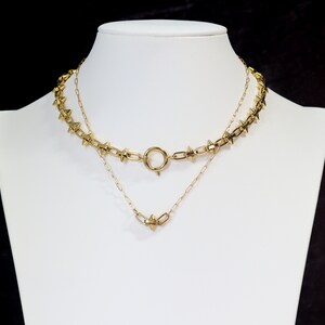 May include: Two gold necklaces, one with a delicate chain and the other with a thicker chain with small spikes. The necklaces are layered on a white mannequin.