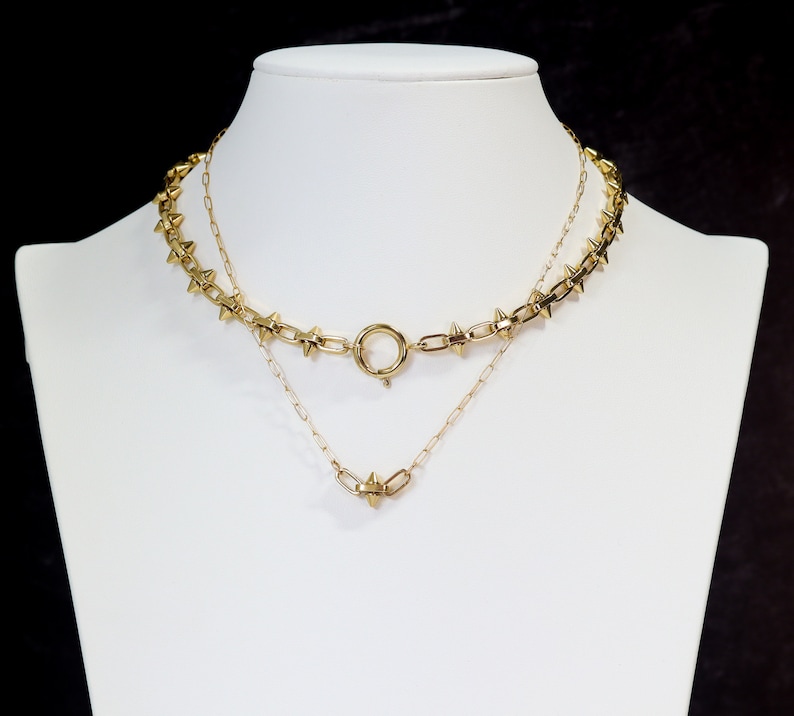 May include: Two gold necklaces, one with a delicate chain and the other with a thicker chain with small spikes. The necklaces are layered on a white mannequin.