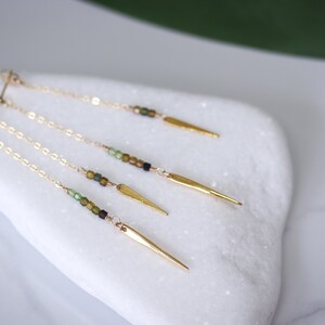 May include: Three pairs of gold dangle earrings with a chain and a gold spike at the end. Each chain has green and brown beads.