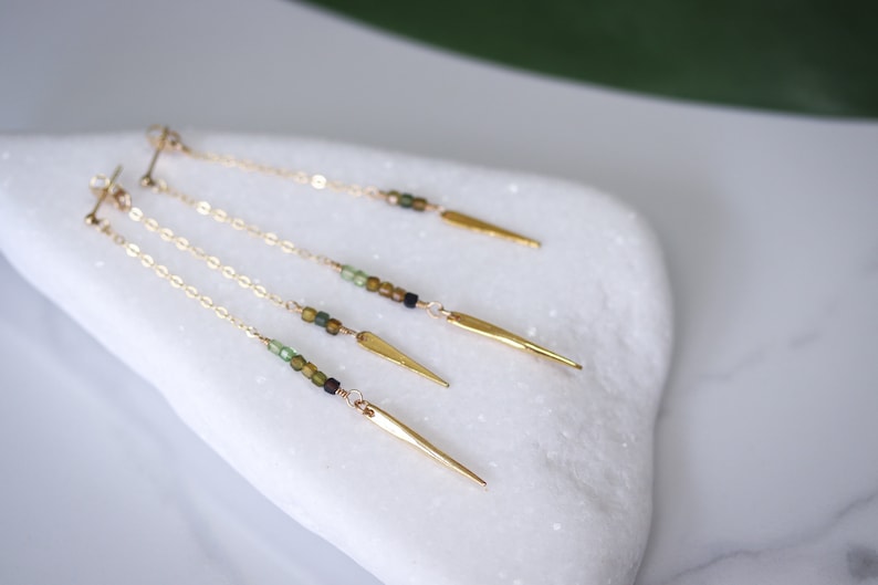 May include: Three pairs of gold dangle earrings with a chain and a gold spike at the end. Each chain has green and brown beads.