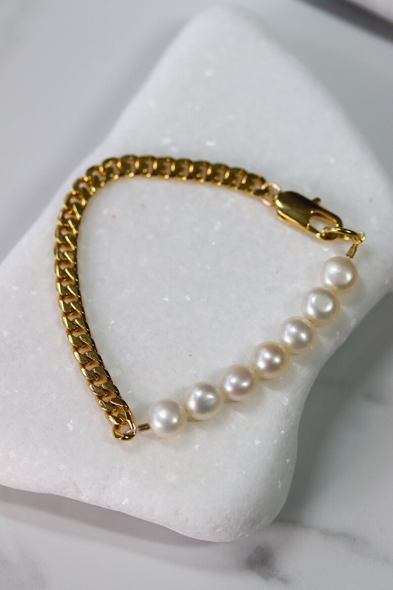 May include: A gold chain bracelet with a row of white pearls. The bracelet has a lobster clasp closure.
