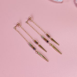 May include: A pair of gold earrings with a delicate chain and small brown, green, and black beads. Each earring has a gold spike at the end.