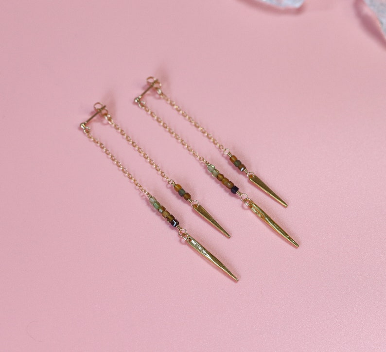 May include: A pair of gold earrings with a delicate chain and small brown, green, and black beads. Each earring has a gold spike at the end.