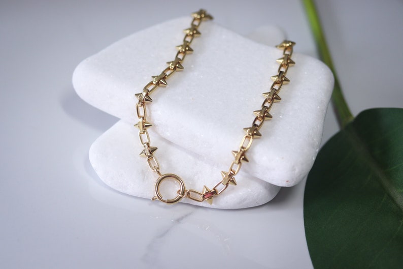 May include: A gold chain necklace with star-shaped spikes. The necklace is on a white surface.