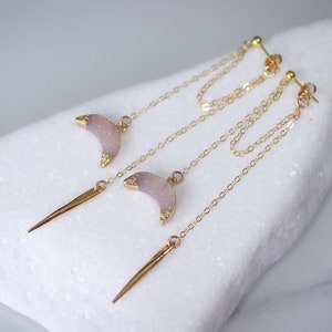 Puede incluir: Un par de pendientes colgantes de oro con dijes de media luna rosa y púas doradas que cuelgan de la parte inferior. Los pendientes están sobre una superficie blanca.